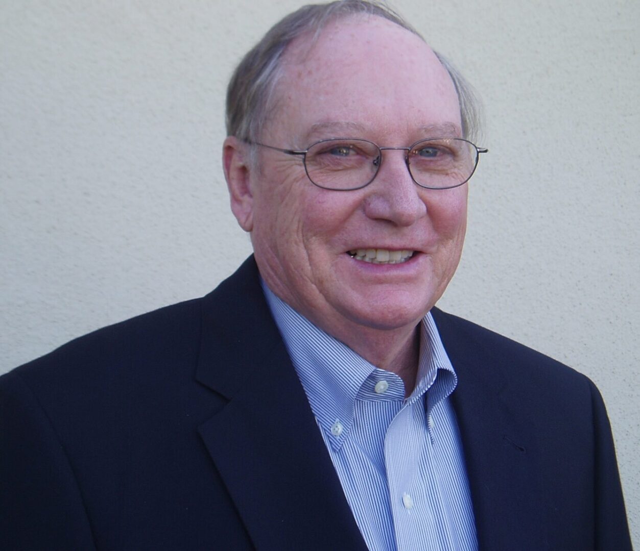 A man in glasses and a suit standing next to a wall.